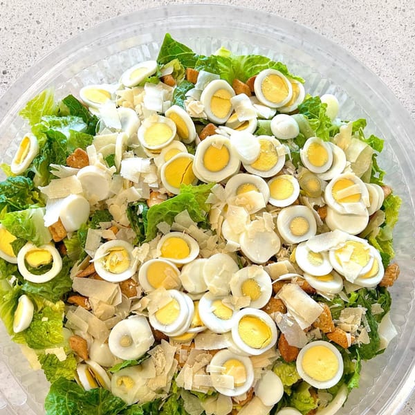 Caesar salad with hard-boiled quail eggs, parmesan shavings, and croutons in a clear container.